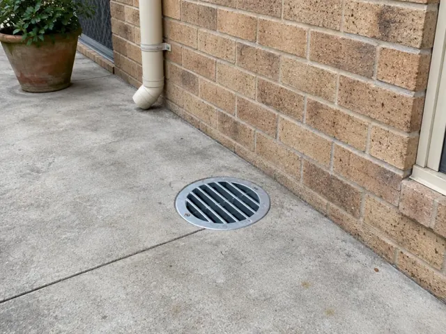 Circular outdoor drain grate set into dry concrete beside a brick wall and potted plant in Engadine